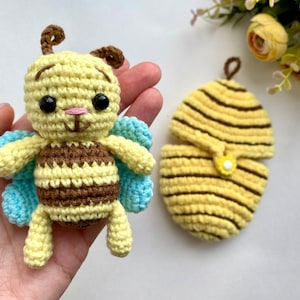 May include: A handmade crochet bee and a matching hive. The bee is yellow, brown, and blue, with black eyes and a pink nose. The hive is yellow with brown stripes and a small yellow flower. The bee measures approximately 3 inches tall.