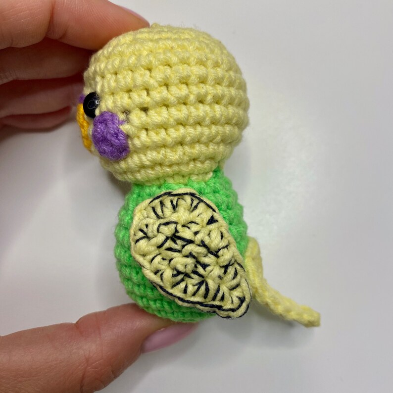 May include: A handmade crochet parakeet in shades of yellow and green. The bird has a yellow head, a green body, and a yellow tail. The wings are green with black detailing, and it has a purple cheek patch and black eye.