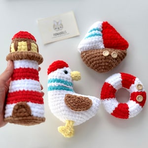 Puede incluir: Conjunto de cuatro juguetes de ganchillo de temática náutica. Un faro a rayas rojas y blancas, una gaviota blanca con rayas azules, un barco a rayas rojas y blancas y un salvavidas a rayas rojas y blancas.