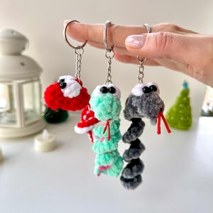 May include: Three crocheted snake keychains with different colored yarn. The snakes are red, green, and gray. Each snake has a black eye and a red tongue.