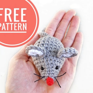 May include: A gray crocheted mouse with white ears and a red nose. The mouse is being held in a hand.