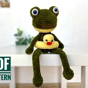 May include: A green crocheted frog amigurumi toy holding a yellow crocheted rubber ducky in a yellow crocheted life preserver. The frog is sitting on a white surface.  PDF PATTERN.