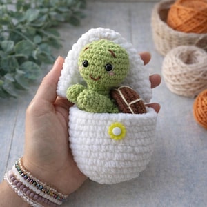 May include: A handmade crochet turtle in a white egg-shaped cozy. The turtle is green with a brown shell and black eyes. The cozy has a yellow flower accent. The turtle is approximately 3 inches tall.