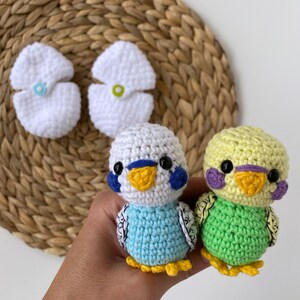 May include: Crocheted parakeet toys in blue and green, with white and yellow accents. The toys are handmade and feature detailed stitching, black eyes, and small yellow feet. Two white crocheted flower-shaped items are also visible.