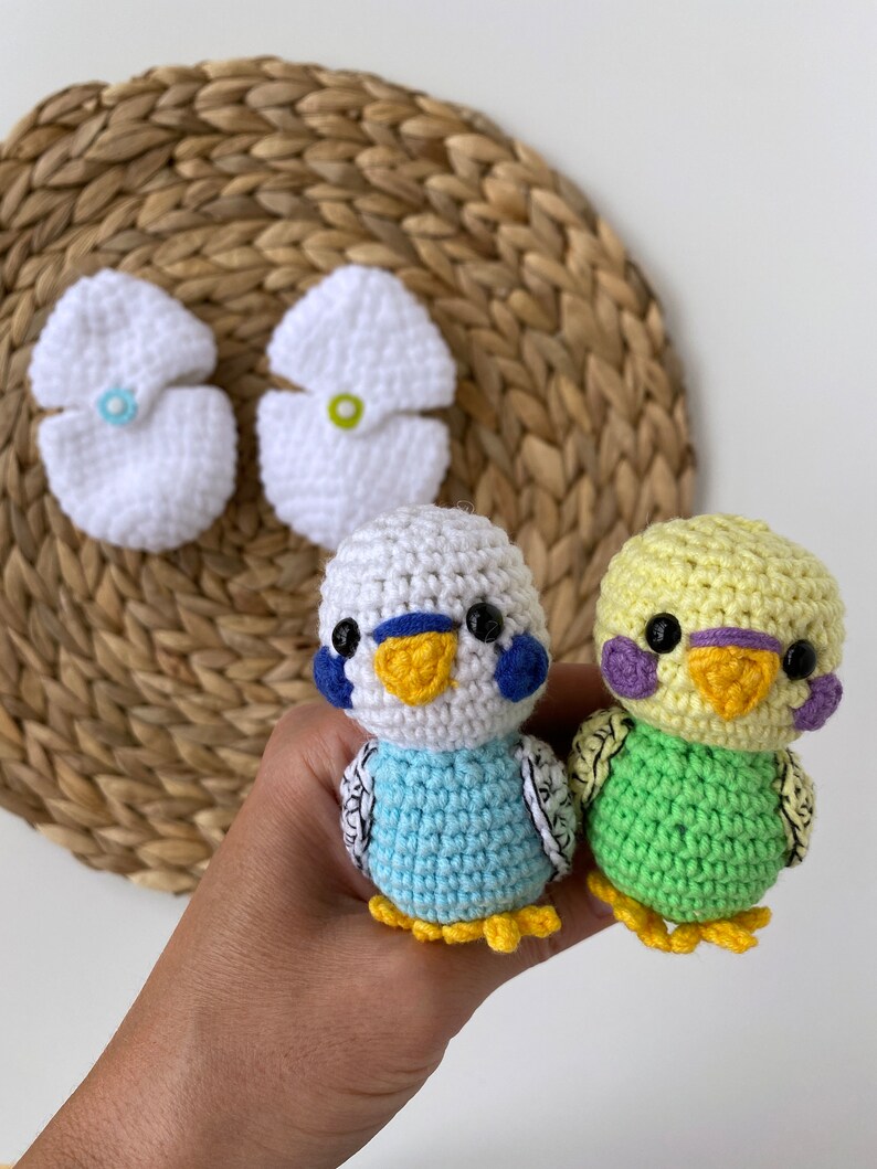 May include: Crocheted parakeet toys in blue and green, with white and yellow accents. The toys are handmade and feature detailed stitching, black eyes, and small yellow feet. Two white crocheted flower-shaped items are also visible.