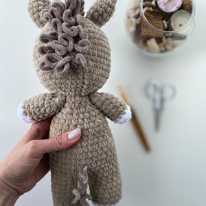 Op de afbeelding: Een gehaakt knuffeldier in de vorm van een paard. Het paard is bruin met een witte manen en staart. Het paard wordt vastgehouden door een hand.
