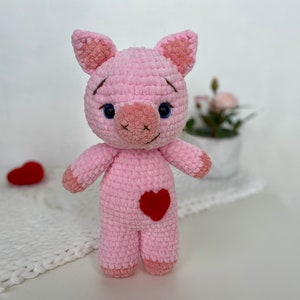 Op de afbeelding: Een roze gehaakt knuffelvarken met een rood hart op zijn borst. Het varken heeft blauwe ogen en een bruine neus.