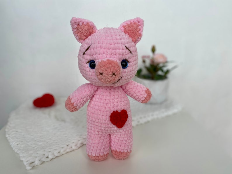 Op de afbeelding: Een roze gehaakt knuffelvarken met een rood hart op zijn borst. Het varken heeft blauwe ogen en een bruine neus.