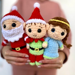 May include: Three crocheted plush toys: a Santa Claus, an elf, and a girl with brown hair. The Santa Claus is wearing a red suit with white trim, a white beard, and black boots. The elf is wearing a green shirt with a black belt, red pants, and a red hat with white trim. The girl is wearing a blue dress with a green trim and a brown headband.