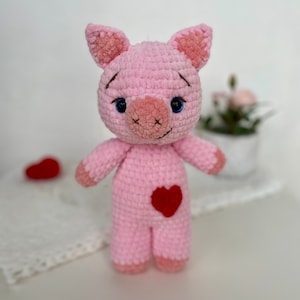 Op de afbeelding: Een roze gehaakt varkens knuffeldier met blauwe ogen en een rood hartje op zijn buik. Het varken staat op een wit oppervlak.