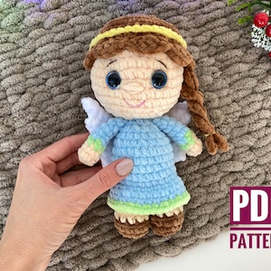 May include: Crocheted angel doll with brown hair, blue dress, and white wings. The doll is holding a yellow flower in its hand. The doll is sitting on a brown textured surface. PDF PATTERN.