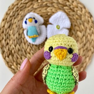 May include: A handmade crochet parakeet in yellow and green, with purple cheek patches and black eyes. The bird has a yellow beak and feet. Two other crochet parakeet figures are in the background, one in white and blue, and the other in white.