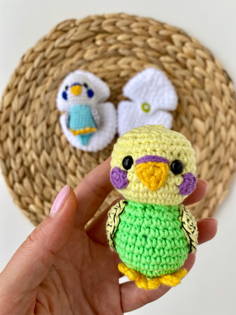 May include: A handmade crochet parakeet in yellow and green, with purple cheek patches and black eyes. The bird has a yellow beak and feet. Two other crochet parakeet figures are in the background, one in white and blue, and the other in white.