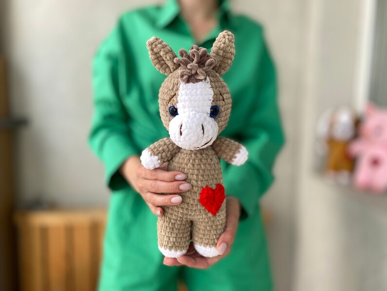 Op de afbeelding: Een gehaakt bruin en wit ezel knuffel met een rood hart op zijn borst. Het speelgoed wordt vastgehouden door een persoon die een groen shirt draagt.