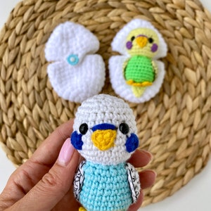 May include: Handmade crochet parakeet with blue, white, yellow, and black details. The parakeet is held in a hand, with two white crochet egg halves and a parakeet hatching from an egg in the background. The parakeet is approximately 3 inches tall.