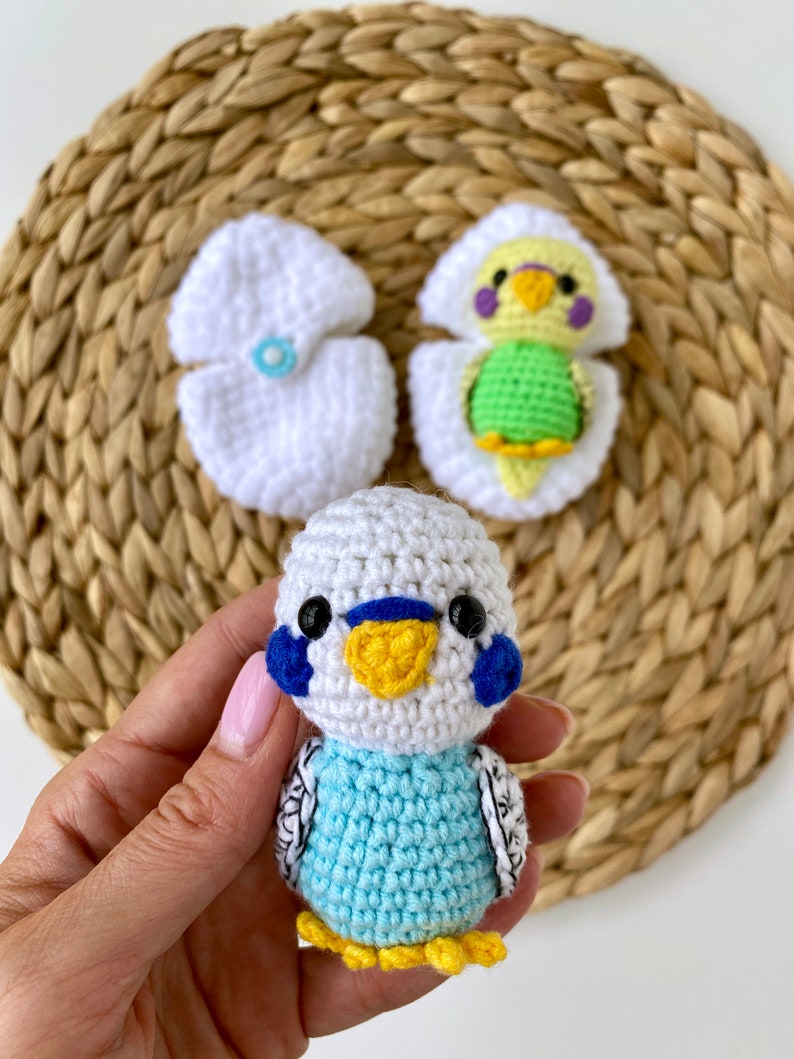 May include: Handmade crochet parakeet with blue, white, yellow, and black details. The parakeet is held in a hand, with two white crochet egg halves and a parakeet hatching from an egg in the background. The parakeet is approximately 3 inches tall.