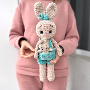 以下が含まれることがあります： 2つの編みぐるみウサギのぬいぐるみ。大きなウサギは小さなウサギを抱っこしています。両方のウサギはベージュで、青いオーバーオールと青いリボンをしています。