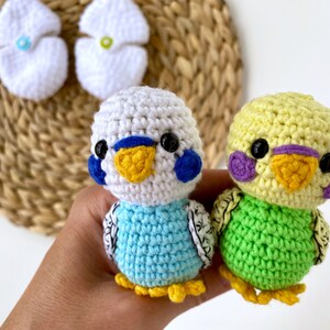 May include: Two crocheted parakeet figurines in the image. One is light blue and white, the other is yellow and green. Both have black eyes, yellow beaks, and detailed wings. Two white crocheted egg-shaped items are in the background.