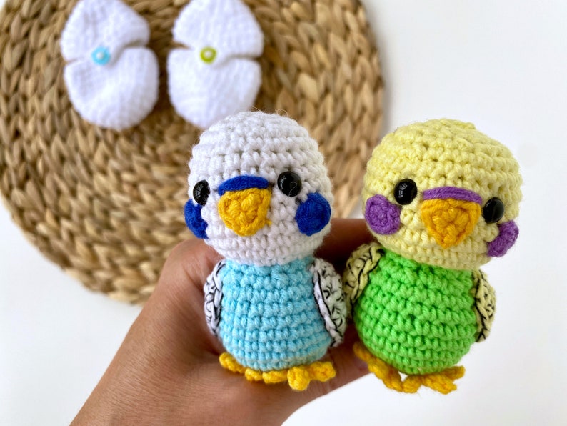 May include: Two crocheted parakeet figurines in the image. One is light blue and white, the other is yellow and green. Both have black eyes, yellow beaks, and detailed wings. Two white crocheted egg-shaped items are in the background.