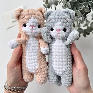 Puede incluir: Dos peluches de gato de ganchillo hechos a mano. Uno es melocotón y blanco, el otro gris y blanco. Ambos tienen ojos negros y narices rosas. Los peluches son sostenidos por manos, mostrando su suave construcción de hilo texturizado. Los gatos tienen orejas pequeñas y brazos extendidos.