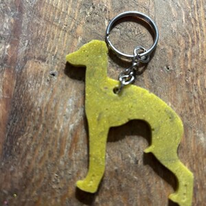May include: A yellow glitter dog-shaped keyring attached to a silver metal ring. The dog's silhouette suggests a slender breed, such as a Greyhound. The keyring has a solid yellow colour with a textured, glittery finish.