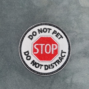 May include: A round patch with a white background and black border. The patch features the text "DO NOT PET" and "DO NOT DISTRACT" in black, surrounding a red stop sign with the word "STOP" in white.