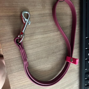 Puede incluir: Una correa de cuero burdeos para perros con un cierre de metal plateado y un acento rosa. La correa está en bucle yace sobre una superficie de madera, junto a un teclado negro.
