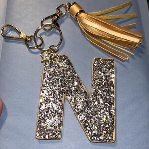 May include: A gold-colored keychain with a glitter-covered letter "N" pendant. The pendant is outlined in a light brown color and is attached to a gold-colored clasp and key ring. A gold tassel hangs from the top of the keychain.