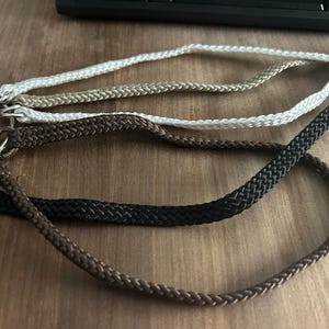 Może przedstawiać: Zbliżenie kilku plecionych smyczy w różnych kolorach: białym, beżowym, brązowym, czarnym i ciemnobrązowym. Każda smycz ma metalowe zapięcie. Smycze leżą na drewnianej powierzchni.