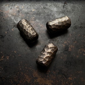 May include: Three hammered brass beads with a rough, textured surface. The beads are a dark gold colour and are arranged in a triangular pattern on a dark grey background.