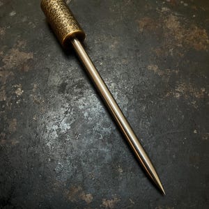 Könnte beinhalten: Eine Metallahle mit einem zylindrischen Griff in strukturierter, gehämmerter Messingoptik und einer langen, glatten, spitz zulaufenden Stahlspitze. Das Werkzeug zeichnet sich durch einen deutlichen Übergang zwischen dem Griff und der Spitze aus.