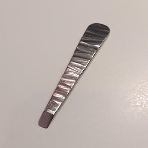 Può includere: Uno scalpello in metallo forgiato a mano con finitura martellata e strutturata. Lo scalpello ha una forma rettangolare che si assottiglia verso un bordo smussato piatto. Il metallo è di colore argento.