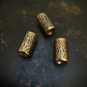May include: Three cylindrical beads with a textured, gold and black finish. The beads have a metallic sheen and a detailed, irregular pattern. The ends of the beads are a solid gold colour. The beads are arranged on a dark, mottled surface.