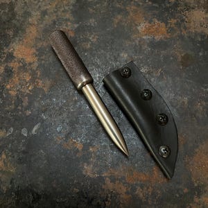 Puede incluir: Un punzón de metal con un mango cilíndrico de agarre moleteado texturizado y una punta cónica lisa. Junto a la herramienta hay una funda negra asegurada con cuatro tornillos de metal oscuro a lo largo de su borde curvo para guardarla.