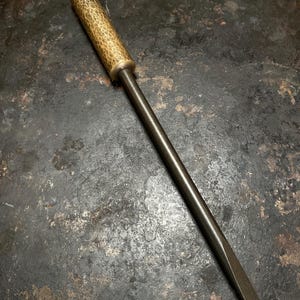 Puede incluir: Un destornillador de metal con un eje de acero cilíndrico, largo y oscuro, con una punta plana y un mango cilíndrico con un acabado dorado martillado y texturizado. La herramienta está colocada en diagonal, mostrando el contraste entre el eje de metal oscuro y el mango metálico con patrón.
