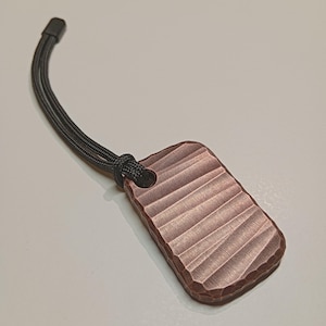 Könnte beinhalten: Ein kupferfarbener, rechteckiger Anhänger mit strukturierter, geriffelter Oberfläche. Eine schwarze Kordel ist durch ein Loch oben befestigt, mit einem schwarzen Kunststoffende. Der Anhänger ist wahrscheinlich ein Schlüsselanhänger oder ein Anhänger.