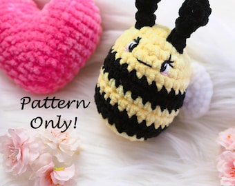 May include: A crocheted bee with black and yellow stripes, black antennae, and a white tail. A pink heart and pink flowers are also in the image. The text "Pattern Only!" is displayed.