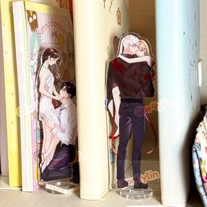 以下が含まれることがあります： アクリル製のアニメキャラクターのスタンドで、ロマンチックなカップルが描かれています。スタンドは本棚の本の間に配置されています。片方は女性の前にひざまずく男性、もう片方は女性を抱きかかえる男性です。スタンドは透明なベースにあります。