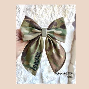 May include: A camouflage print bow with the name "Zamora" embroidered in black. The bow has a khaki-colored center and is from "the Awkward Little turtle". The bow is likely for a pet.