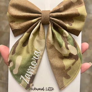 Peut inclure: Un grand n&oelig;ud papillon &agrave; motif camouflage avec le nom "Zamora" brod&eacute; en &eacute;criture blanche. Le n&oelig;ud est fait d'un tissu textur&eacute; et est pr&eacute;sent&eacute; sur une carte blanche. Le texte "the Awkward Little turtle" est visible.