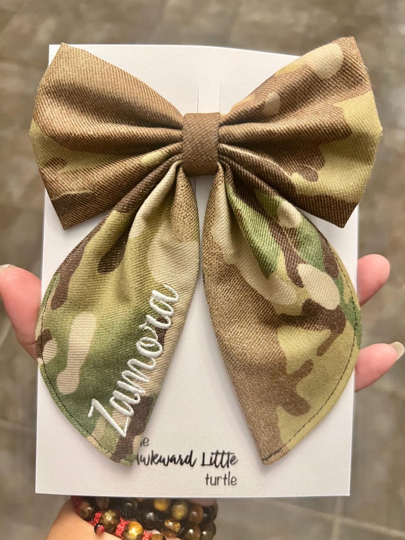 Peut inclure: Un grand n&oelig;ud papillon &agrave; motif camouflage avec le nom "Zamora" brod&eacute; en &eacute;criture blanche. Le n&oelig;ud est fait d'un tissu textur&eacute; et est pr&eacute;sent&eacute; sur une carte blanche. Le texte "the Awkward Little turtle" est visible.