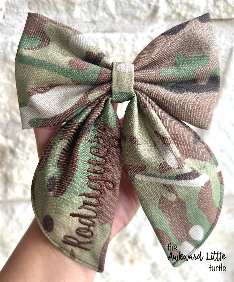 Peut inclure: N&oelig;ud papillon &agrave; motif camouflage vert, marron et beige. Le nom "Rodriguez" est brod&eacute; en fil marron. Le texte "the Awkward Little turtle" est imprim&eacute; en noir en bas &agrave; droite.