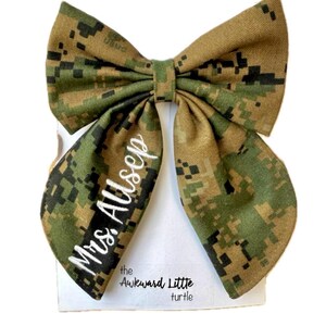 Peut inclure: Un n&oelig;ud papillon de grande taille avec un motif camouflage vert, marron et beige. Le texte "Mrs. Allsep" est brod&eacute; en blanc sur un c&ocirc;t&eacute;. Le n&oelig;ud est pr&eacute;sent&eacute; sur une carte blanche avec le texte "the Awkward Little turtle".