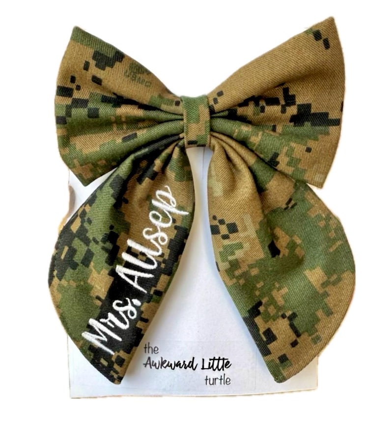 Peut inclure: Un n&oelig;ud papillon de grande taille avec un motif camouflage vert, marron et beige. Le texte "Mrs. Allsep" est brod&eacute; en blanc sur un c&ocirc;t&eacute;. Le n&oelig;ud est pr&eacute;sent&eacute; sur une carte blanche avec le texte "the Awkward Little turtle".