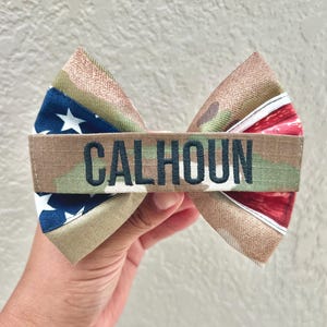 May include: A handmade bow tie featuring a camouflage pattern with an American flag design on one side and red stripes on the other. The name "CALHOUN" is embroidered in black across the center. The bow tie is held in a hand.