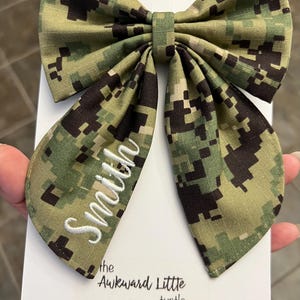 Peut inclure: Un grand n&oelig;ud papillon &agrave; motif camouflage, avec le nom "Smith" brod&eacute; en blanc. Le n&oelig;ud est pos&eacute; sur une carte blanche avec le texte "the Awkward Little turtle".