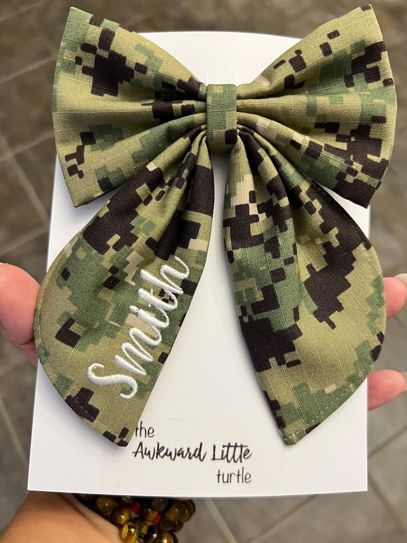 Peut inclure: Un grand n&oelig;ud papillon &agrave; motif camouflage, avec le nom "Smith" brod&eacute; en blanc. Le n&oelig;ud est pos&eacute; sur une carte blanche avec le texte "the Awkward Little turtle".