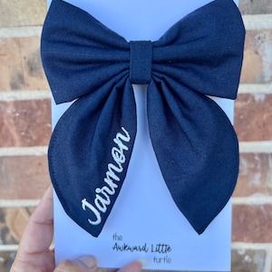 Peut inclure: Un n&oelig;ud papillon bleu marine avec le nom "Jarmen" brod&eacute; en &eacute;criture blanche. Le n&oelig;ud est pos&eacute; sur une carte blanche avec le texte "the Awkward Little turtle" en noir.