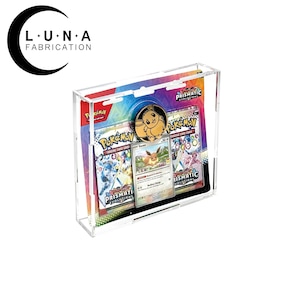 May include: A clear acrylic display case with a Pokemon card collection inside. The case features a holographic Pokemon card with a Eevee evolution, a gold coin with a Eevee image, and a second Pokemon card with a Eevee evolution. The case has the text "Prismatic Evolutions" on the front.