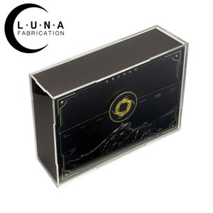 May include: A rectangular display case with a black and gold design. The case has a clear acrylic front and sides, with the text "LUNA FABRICATION" and a crescent moon logo in the upper left corner. The front panel features a gold emblem and text.